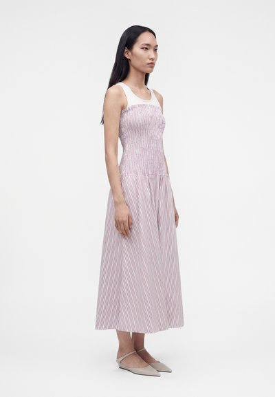 Γυναίκα στέκεται, φοράει αμάνικο midi φόρεμα ροζ και λευκές ριγέ γραμμές και μπεζ μυτερές μπαλαρίνες, με φόντο απλό και φωτεινό.