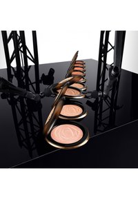 Zehn kompakte Rouge mit einem Rosenmuster, jeweils in einem runden schwarzen Etui mit goldenen Akzenten, angeordnet auf einer glänzend schwarzen Oberfläche.