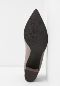 Chaussure à talon haut marron avec un bout pointu. La semelle est noire avec des motifs de pièces en relief et le nom de la marque "KAISER" affiché de manière visible.