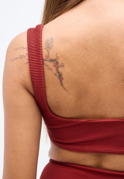 Ερυθρόριγος κορυφή καλλιέργειας από ύφασμα με πλατιά τιράντα, που αποκαλύπτει τον τόνο του δέρματος και ένα λουλουδένιο τατουάζ στον ώμο και στην άνω πλάτη.