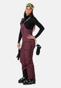 Maronfarvet ski-overall med sorte lynlåse og lommer, bæres over et sort basislag. Sorte skihandsker og støvler fuldender outfittet.