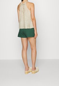 Haut en tissu beige à encolure halter avec un lien au cou, associé à un short vert et des sabots en bois clair avec un talon surélevé.
