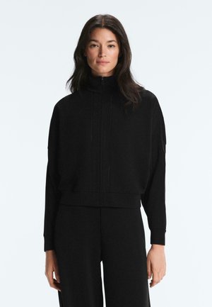 Frau mit langen dunklen Haaren, die eine schwarze Reißverschlussjacke mit hohem Kragen und passende schwarze Hose trägt, steht vor einem einfarbigen weißen Hintergrund.