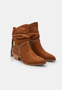 Botines de ante en marrón, con un diseño holgado, puntera afilada, tacón de madera y tachuelas decorativas en azul y plata a lo largo de los lados.