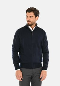 Maglione zip-up blu navy con colletto ufficiale, polsini e vita a costine. Texture morbida e design sinuoso; indossato sopra una camicia con colletto.