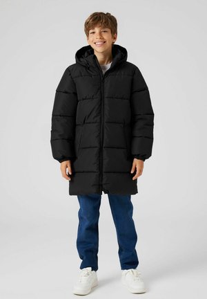 Cappotto a collo alto di colore nero, con design trapuntato e polsini elastici. Abbinato a jeans blu e sneakers bianche.