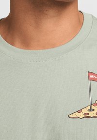 Detailní záběr na krk a horní část hrudníku, oblečený v světle zelen�é košili s kresleným kusem pizzy a červenou golfovou vlajkou na pravé straně.