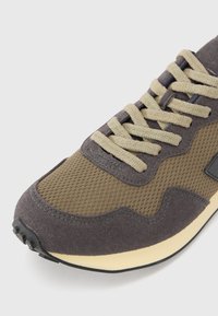 Adidaș maro din suede și plasă, cu șirete bej, suprafață texturată și talpă din cauciuc negru. Prezintă un design stratificat și accente de cusătură verzi.