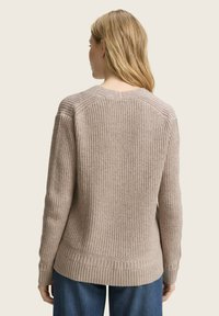 Woman with long blonde hair wearing a beige chunky knit sweater and blue jeans, facing away against a plain light background.