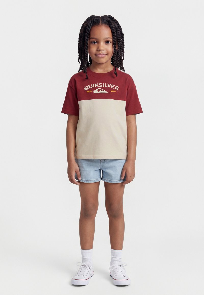 Νεαρή κοπέλα με πλεξούδες, φορώντας κόκκινη και μπεζ μπλούζα Quiksilver, τζιν σορτς, λευκές κάλτσες και λευκά αθλητικά παπούτσια, όρθια.