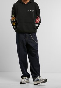 Osoba v černé mikině s barevnými nášivkami na rukávech, tmavě modrých džínách s řetězovým doplňkem a stříbrných běžeckých botách na šedém pozadí.