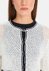 White knitted cardigan with a textured open weave, navy trim, and five black buttons. Features a round neckline and contrasting striped detail.