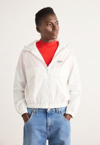 White lightweight zip-up jacket with a hood and elastic cuffs, featuring a chest pocket and subtle logo. Paired with a red turtleneck and blue jeans.