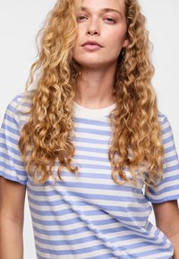 Noor inimene, kellel on pikad lokkis heledad juuksed, kandes hele sinise ja valge triibulise lühikeste varrukatega särgi, vaadates otse ette neutraalse ilmega.