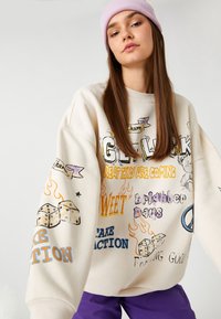 Sudadera color crema con estampados gráficos, que presenta texto e ilustraciones en varios colores. Corte holgado, textura suave, puños acanalados.