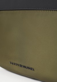 Olivgrüne Nylon-Tasche mit einer strukturierten schwarzen Oberseite, die sichtbare Nähte aufweist und ein eingeprägtes "Tommy Hilfiger"-Logo auf der Vorderseite trägt.