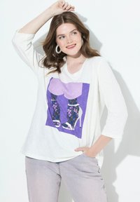 Valkoinen kolme neljännespuolen hihainen pusero, jossa on graafinen printti jaloista kukkakuosisissa legginsseissä ja korkokengissä purppuran taustan edessä.