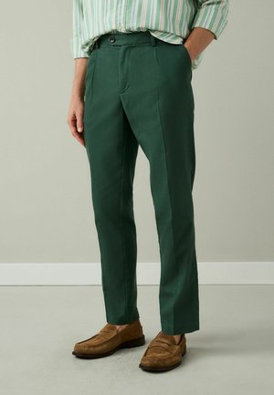 Mann trägt dunkelgrüne maßgeschneiderte Hose, hellbraune Loafers und ein grün-weiß gestreiftes Hemd, eine Hand in der Tasche.