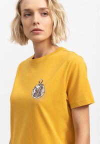 T-shirt di cotone gialla con una grafica di un orologio da tasca sul davanti. Collo rotondo e maniche corte con una texture liscia.