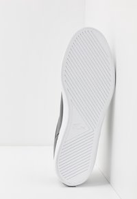 Semelle d'une sneaker blanche avec des rainures diagonales et un petit logo crocodile embossé au centre, inclinée contre un mur d'angle blanc.