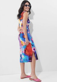 Figurbetontes, ärmelloses Kleid im mehrfarbigen abstrakten Druck mit einem Seitenschlitz. Kombiniert mit pinken, strukturierten Slides und blauen Sonnenbrillen.