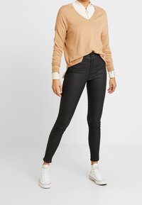 Beige V-kauluksinen neuletakki, löysä malli ribbauskumilla, valkoisen paidan päällä. Mustat slim fit -housut ja valkoiset lenkkarit täydentävät asun.