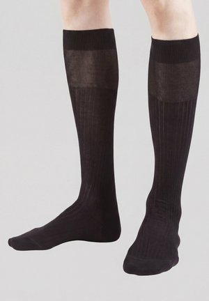 Benen met zwarte, geribbelde kniekousen met verstevigde bovenkanten tegen een effen lichte achtergrond.