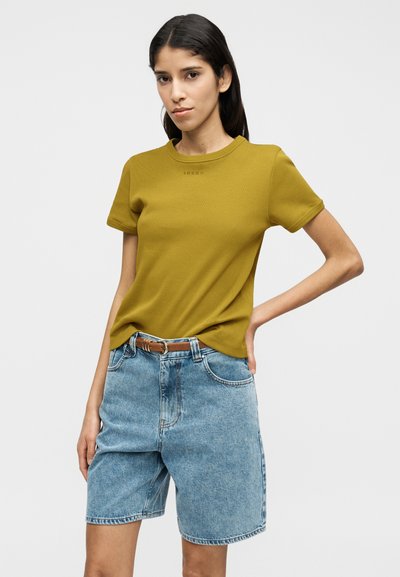 Μουσταρδί κίτρινο κοντομάνικο μπλουζάκι με στρογγυλή λαιμόκοψη και μικρό λογότυπο, συνδυασμένο με ανοιχτόχρωμο τζιν σορτς και καφέ ζώνη.