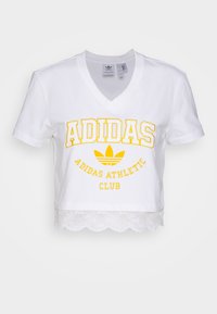 Baltas trumpas marškinėliai su V formos iškirpte, su geltona "ADIDAS" tekstu ir logo bei nėriniais apačioje. Pagaminta iš medvilnės.