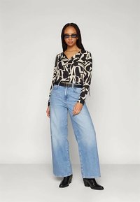 Γυναίκα με πλεξούδες που φορά μαύρα γυαλιά ηλίου, αφηρημένο λευκό και μαύρο μακρυμάνικο μπλουζάκι, ανοιχτόχρωμα jeans με φαρδιά ποδιά, μαύρη ζώνη και μαύρες μπότες.