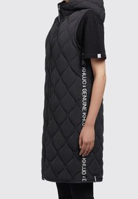 Person wearing a sleeveless black quilted vest with a front zipper and printed white text on the sides, over a black short-sleeve shirt.