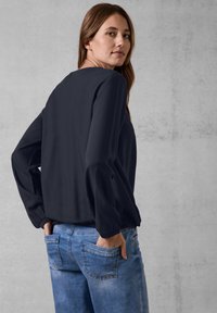 Marineblå langærmet bluse med en afslappet pasform og elastiske manchetter, parret med lyseblå denimbukser med baglommer og en slidte tekstur.
