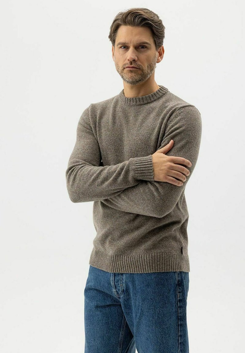 Mann mit kurzen braunen Haaren und Bart, der einen braunen Strickpullover und blaue Jeans trägt, steht mit verschränkten Armen vor einem weißen Hintergrund.