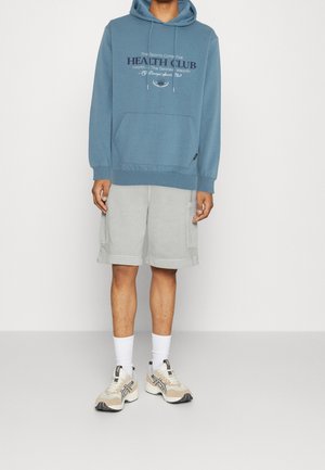 Sudadera azul con capucha y bolsillo frontal, con texto "HEALTH CLUB". Combinada con pantalones cortos gris claro y zapatillas crema.