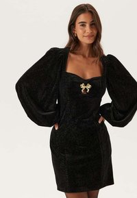 Svart sammetsklänning med stora puffärm, fyrkantig halsringning och en guldbågdetalj vid bröstet. Tyget har ett subtilt mönster med textur.