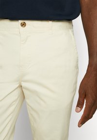 Pantalons en coton beige avec une texture lisse, coupe droite et fermeture à bouton. Le tissu est léger, adapté à une tenue décontractée.