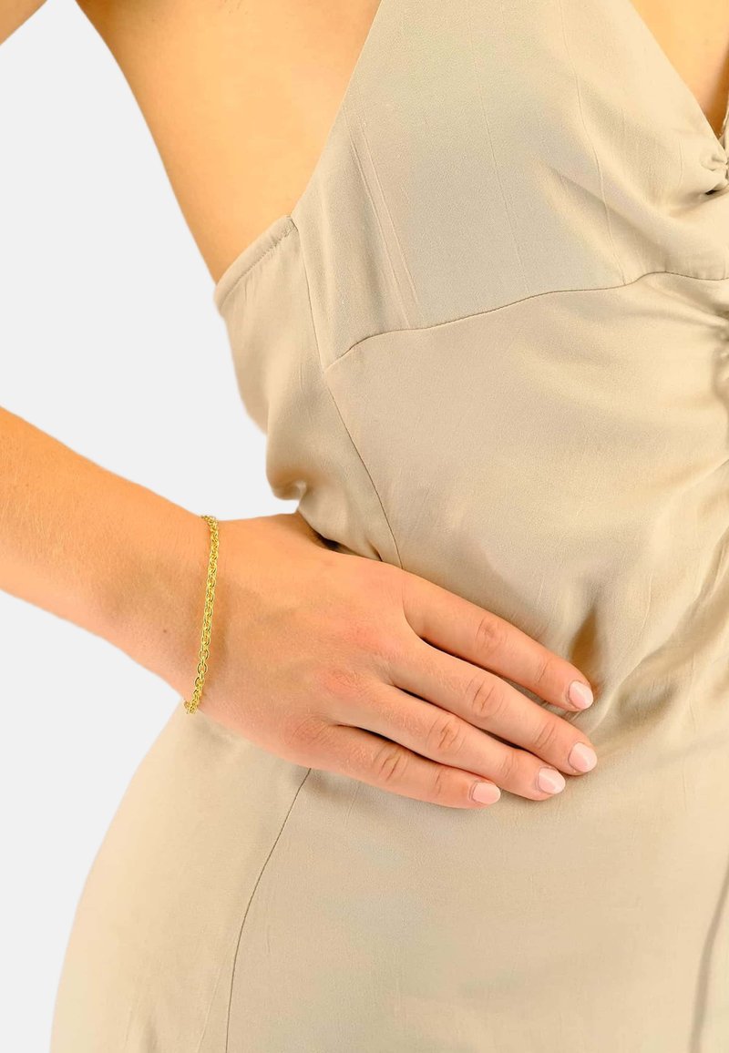 Vêtement beige à texture lisse avec un col drapé. Un bracelet chaîne en or est visible sur une main reposant sur la hanche.