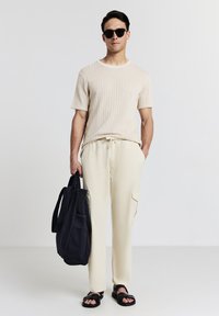 Mann trägt ein beiges, kurzes Strickshirt, helle Hose, schwarze Sandalen und Sonnenbrille und hält mit einer Hand eine große schwarze Tragetasche.