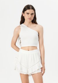 Fehér bordázott crop top egy vállra eső dizájnnal, amit egy réteges fehér fodros mini szoknyával párosítanak, amely finom hímzéssel díszített.