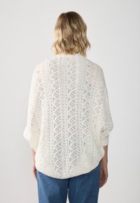 Bílý pletený svetr s volným střihem, s otevřeným vzorem, širokými rukávy a žebrovanými manžetami. Nosený přes modré džíny.