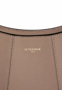 Borsa in pelle di colore taupe chiaro, con finitura texture, logo dorato "LE TANNEUR 1898" e dettagli di cucitura visibili.