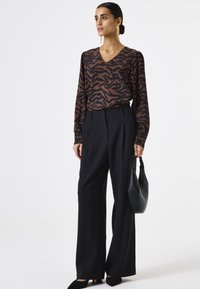 Bruine en zwarte zebra-print blouse met lange mouwen en een V-hals, gecombineerd met hoge zwarte broek. Model houdt een zwarte tas vast.