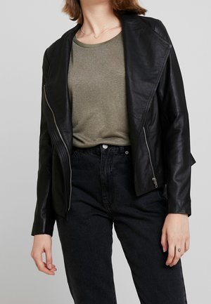 Person trägt eine schwarze Lederjacke über einem olivgrünen T-Shirt und hoch taillierte schwarze Jeans vor einem einfarbigen grauen Hintergrund.