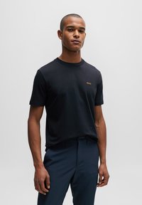 Camiseta negra de manga corta en tejido suave con cuello redondo y un pequeño logo dorado en el pecho, combinada con pantalones azul marino.