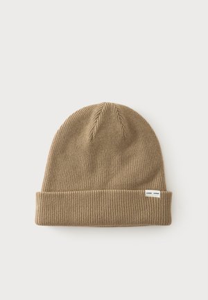 Bonnet en maille brun clair avec un revers plié et une petite étiquette blanche sur le poignet, sur un fond clair uni.
