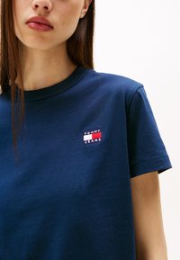 Tengerészkék pamut póló rövid ujjakkal. A bal mellen található egy piros, fehér és kék logó folt, amelyen az "TOMMY JEANS" felirat olvasható.