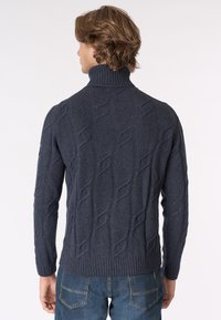 Uomo che indossa un pullover a collo alto in maglia cablè blu navy e jeans blu, mostrato di spalle su uno sfondo bianco semplice.