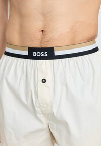 Heren shorts in crèmekleur met een zwarte tailleband waarop een gouden en witte streep staat, en de merknaam "BOSS" duidelijk zichtbaar.