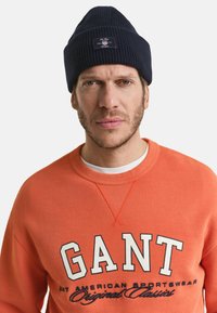 Marine gebreide beanie met logo patch; oranje sweatshirt met witte en donkerblauwe tekst "GANT AMERICAN SPORTSWEAR Original Classic." Geschikt textiel.
