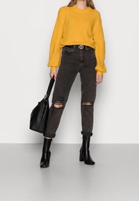 Femme portant un pull jaune, un jean noir déchiré, des bottines noires, tenant un sac à main noir sur fond blanc.